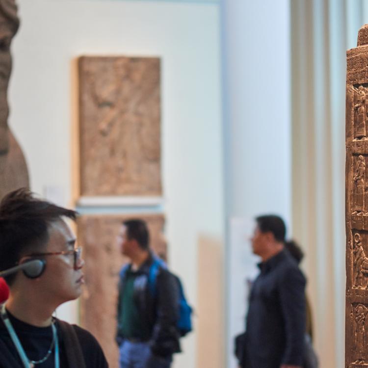 A visitor listening to an audio guide while looking at the Black Obelisk of Shalmaneser III in Room 6 Assyrian Sculpture. In the background, the statue of a human-headed winged lion is visible