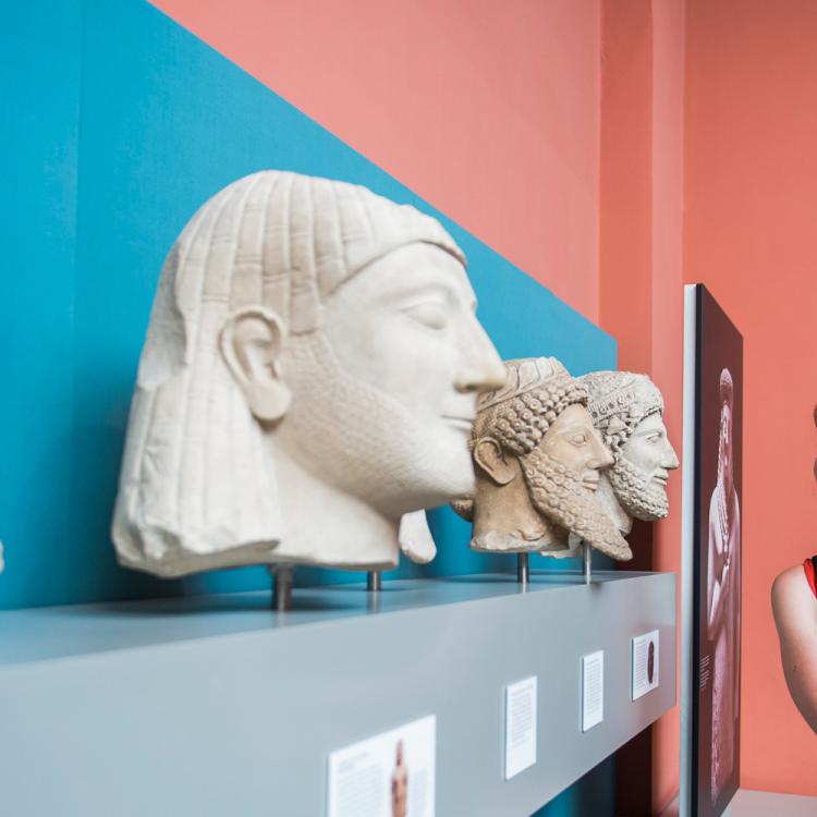 A visitor and her child examining three sculptures of heads. Each statue is adorned with a hat, curly beard and has a broken nose.