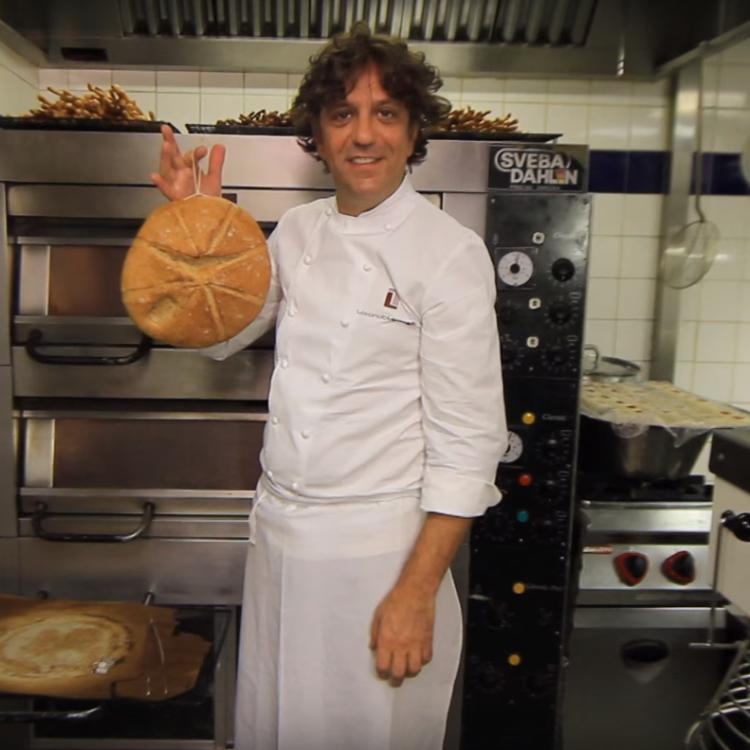 Chef Giorgio Locatelli holds a round loaf of bread in a professional kitchen.