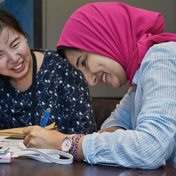 Two women at a desk smiling, one of them writing on paper.