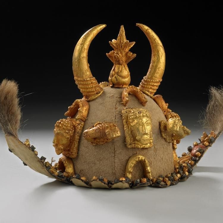 Ceremonial cap made of hide, adorned with gold details, including golden horns, golden badges of faces, and raised flaps with fur at each end