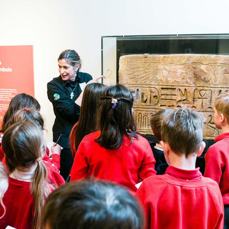 A group of school children watching a museum guide talk about a large hieroglyphs panels