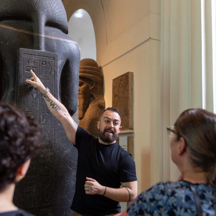 Matthew wears a black tshirt and points at hieroglyphs on a large ancient Egyptian sculpture.
