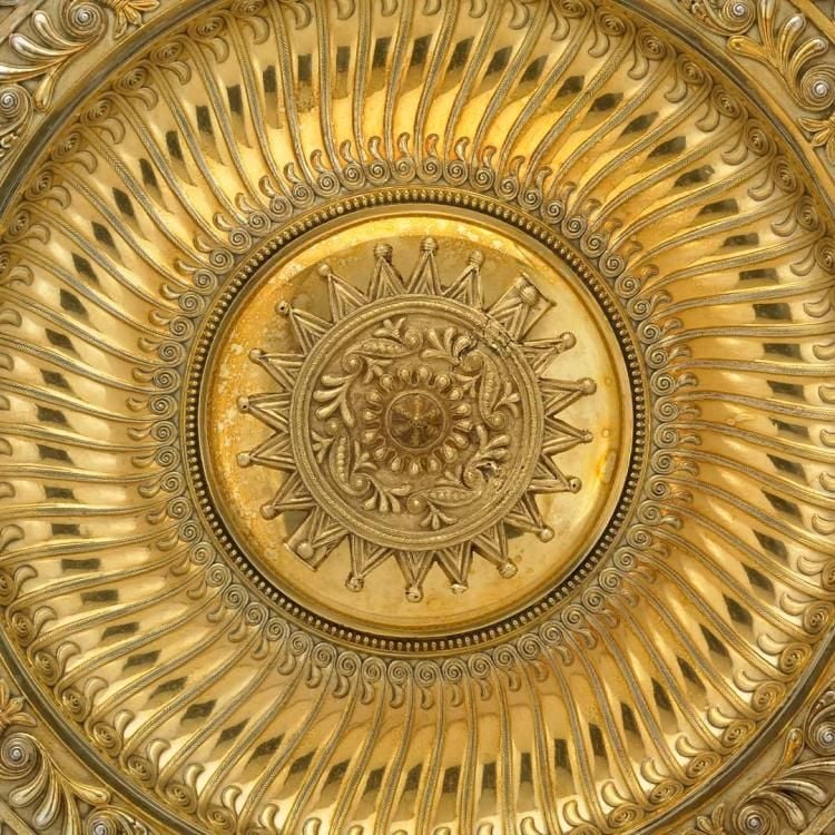 An ornate gold circular plate standing on its edge with a raised circular pendant at its centre on a grey background.
