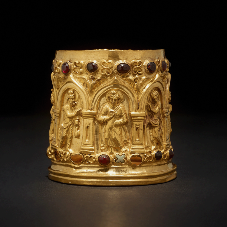 A round gold reliquary container studded with red and orange stones featuring intricate representations of the Buddha flanked by the gods Indra and Brahma.