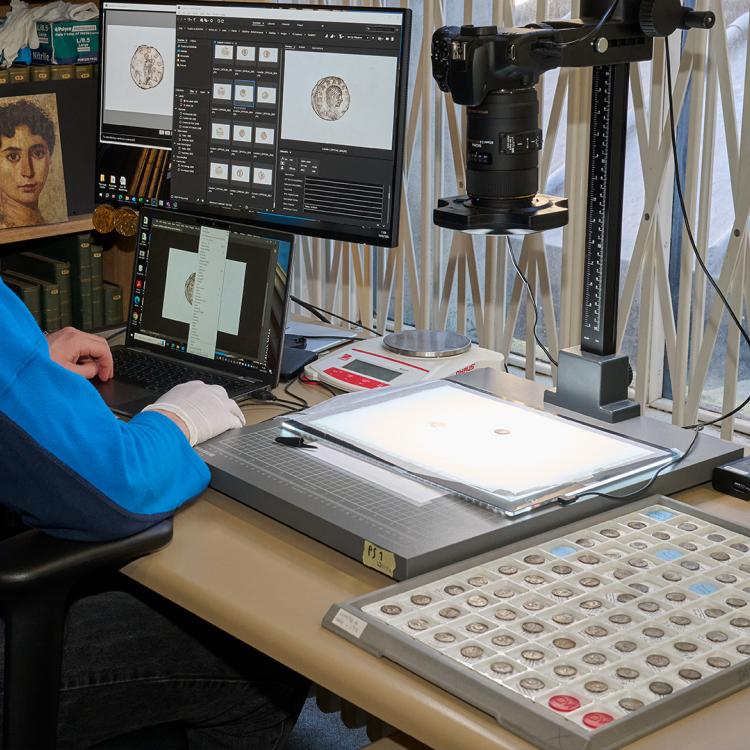 Member of staff working on their computer at a desk, with a tray of coins to one side which are being photographed one-by-one under a mounted camera.