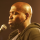 Kareem Parkins-Brown performing with a mic in his right hand and mobile in his left, against a black background. He wears a light-coloured T-shirt and a black unzipped sweatshirt, his head is clean shaven and he has short facial hair.