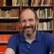 Ian has fair hair and a beard and is wearing a blue collared t-shirt sitting in front of a bookcase.