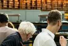 View of several patrons from behind, facing into the Reading Room which is lined with desks and filled bookshelves against the far wall.