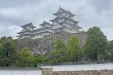 View of a multi-storey building in a traditional Japanese architectural style, atop a high brick wall front by a row of green trees.