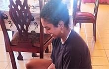 Chrisyl wears a dark t-shirt and sits on a stool in a room with a yellow-tiled floor and a table and chairs