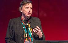 Lissant Bolton wears a colourful scarf and a dark jacket, and speaks into a microphone in front of a red background