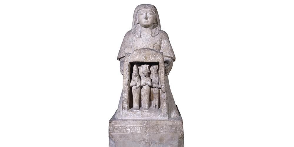 A kneeling statue Panehesy who holds between his hands a naos containing the figures of Horus, Osiris and Isis and surmounted by a winged sun-disk.