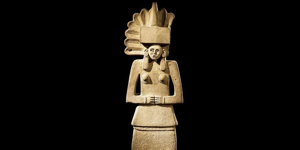 Female figure made of sandstone, standing with hands folded in front and with a fan-shaped headdress.