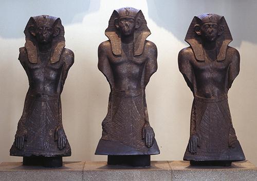 Row of three statues of Senusret III wearing nemes, legs and arms lost but hands still present on aprons.