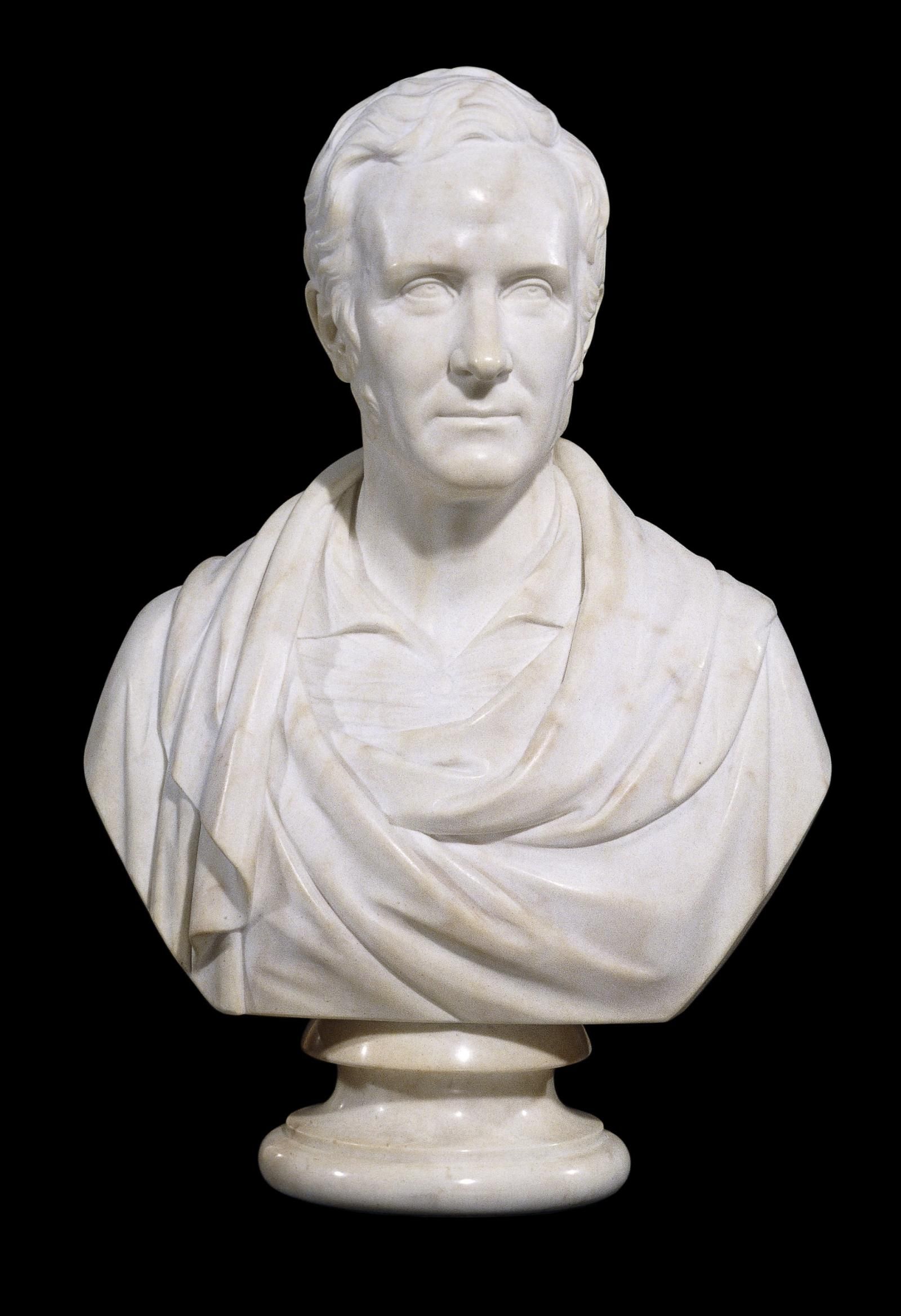 A marble bust of a male figure  wearing a robe in the classical style, attached to a polished marble socle. 