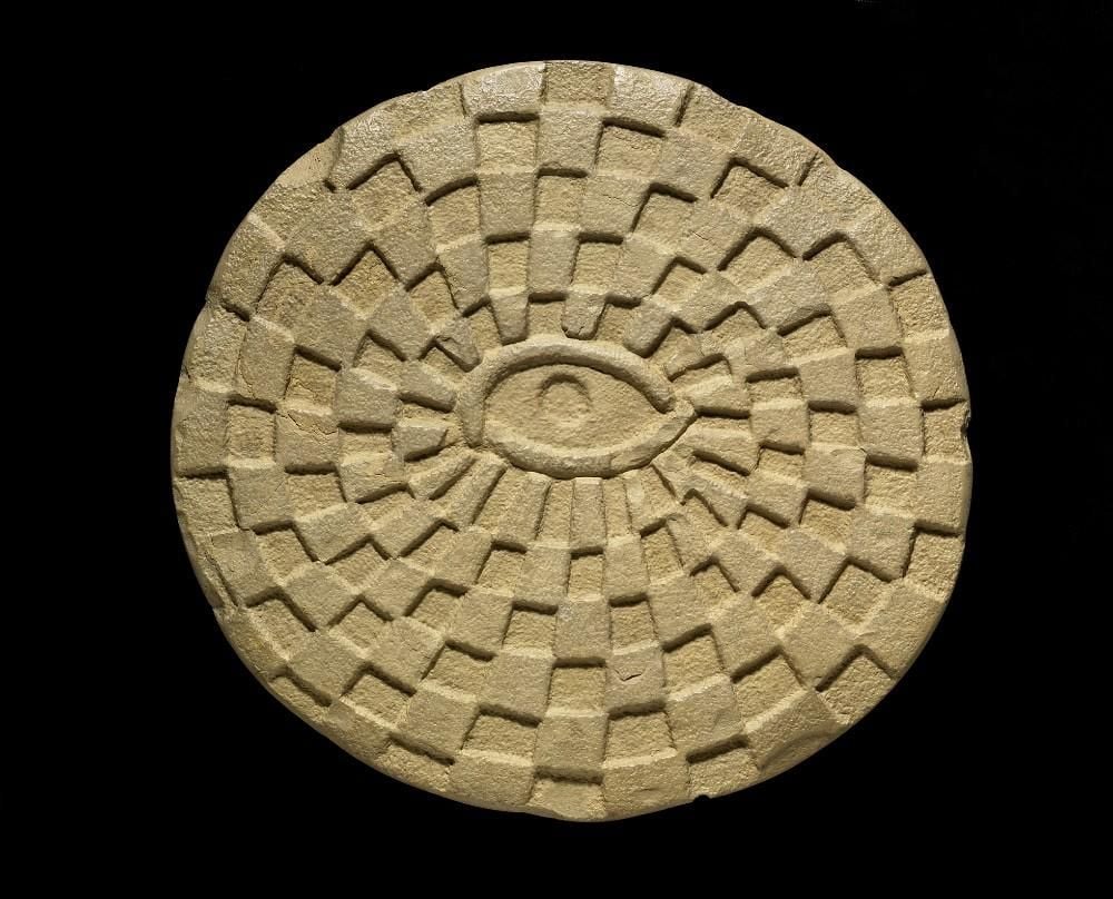 A circular game board of pale yellow limestone. The upper surface is carved with a spiral pattern of square projections and hollows