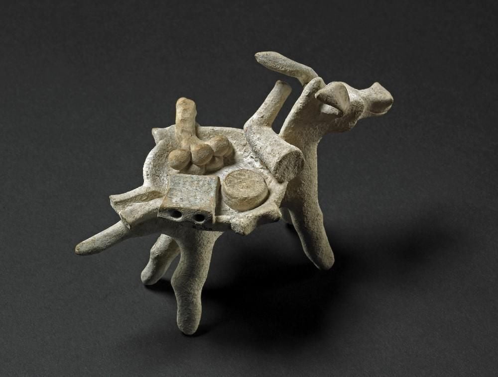 Terracotta figure of a mule carrying a mortarium. Inside it are a pestle, a cheese-grater, a round cheese, and a bunch of garlic.