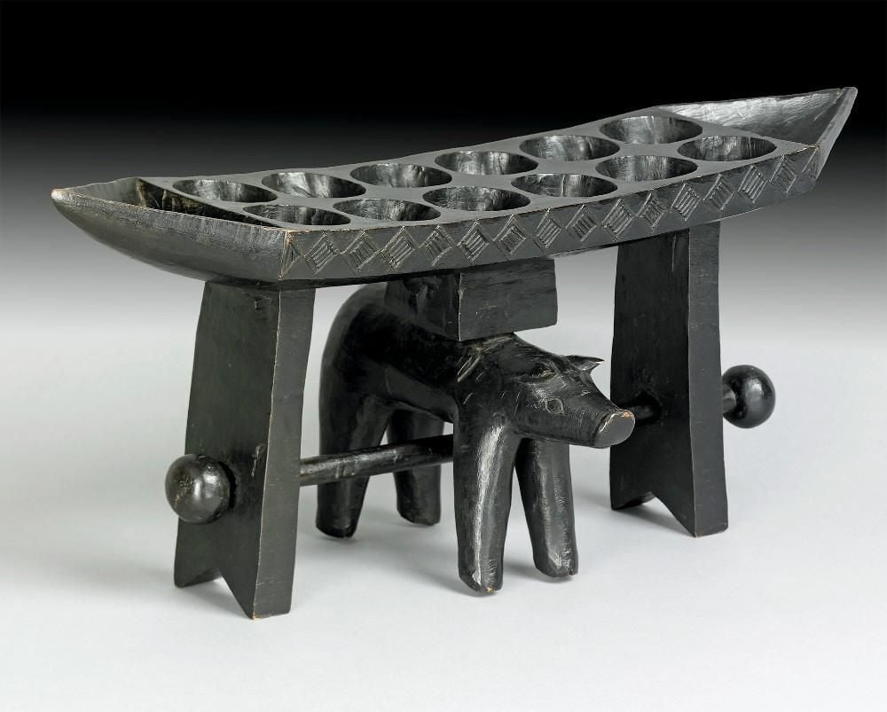  Rectangular board carved with twelve round cavities, pointed ends, on a stand with pig-like animal underneath