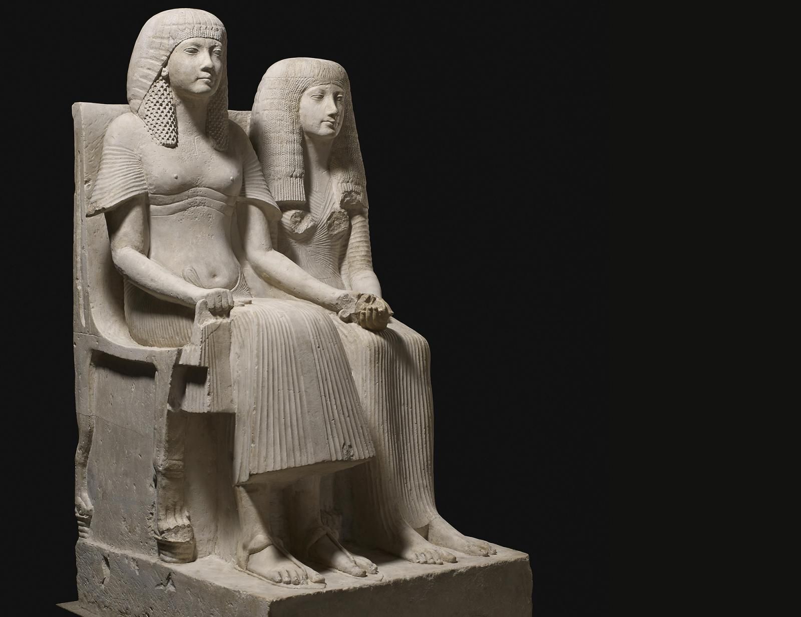 Limestone statue of Horemheb and one of his wives seated on chairs holding hands.