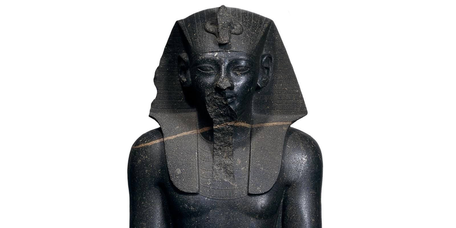 A granite statue of a Egyptian king, upper body from chest to face visable. The figure is wearing a head-dress and there is damage to the sculpture on his chin.