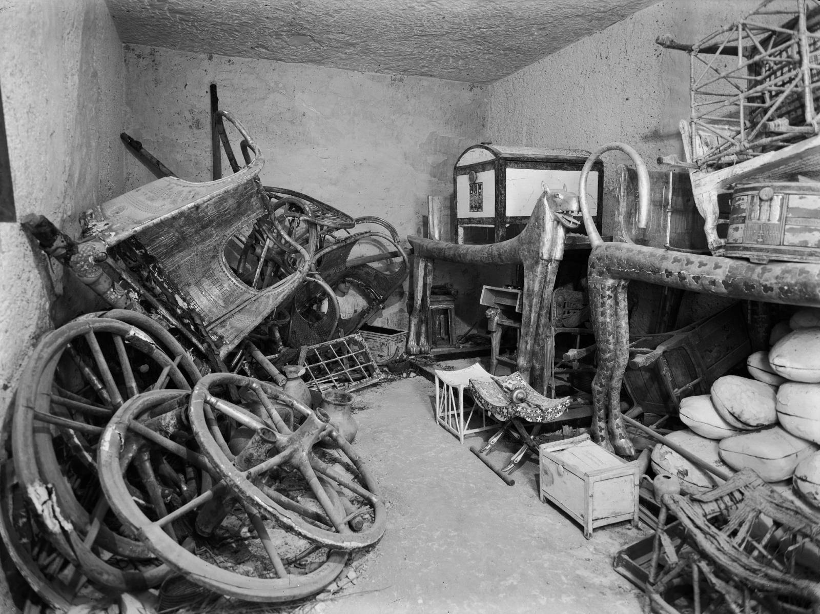 Black and white photo showing the inside of Tutankhamun's tomb. Objects in the photo include cat statues, wheels and boxes.