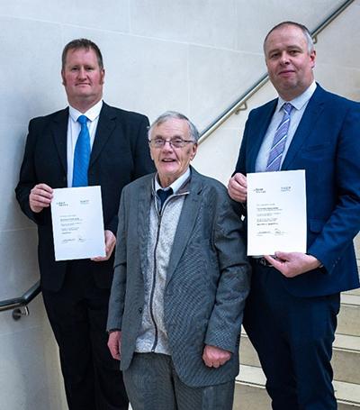 Three men wearing suits standing on a staircase with a metal rail and white wall behind them. Two are holding certficates. 