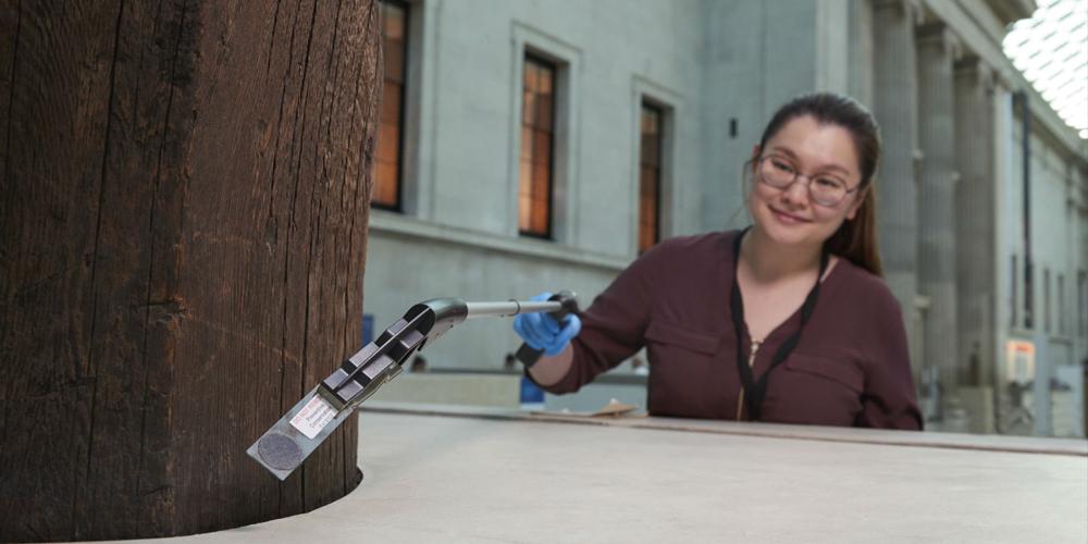 Collection staff member picks up a dust sampling slide from the base of a totem pole in the Great Court using a reach extender tool.
