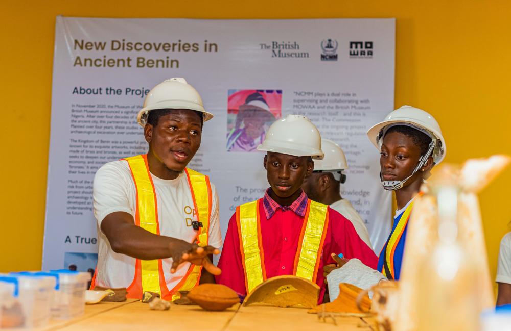 Person talking to two students, explaining objects on a table, all in hard hats and high-visibility sashes, project poster behind