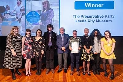 A group of smartly dressed figures stand in front of a large screen with the words "Winner" and "The Preservative Party Leeds City Museum". 