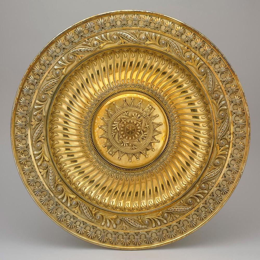 An ornate gold circular plate standing on its edge with a raised circular pendant at its centre on a grey background.