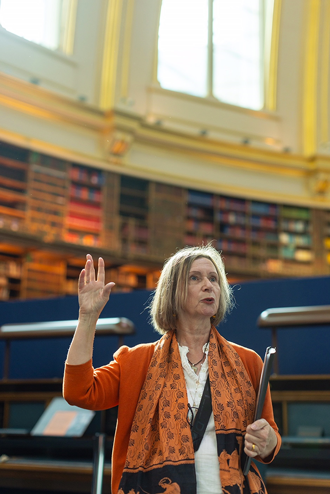 Kathryn wears an orange jacket and scarf, and gestures with one hand towards the domed roof of the reading room