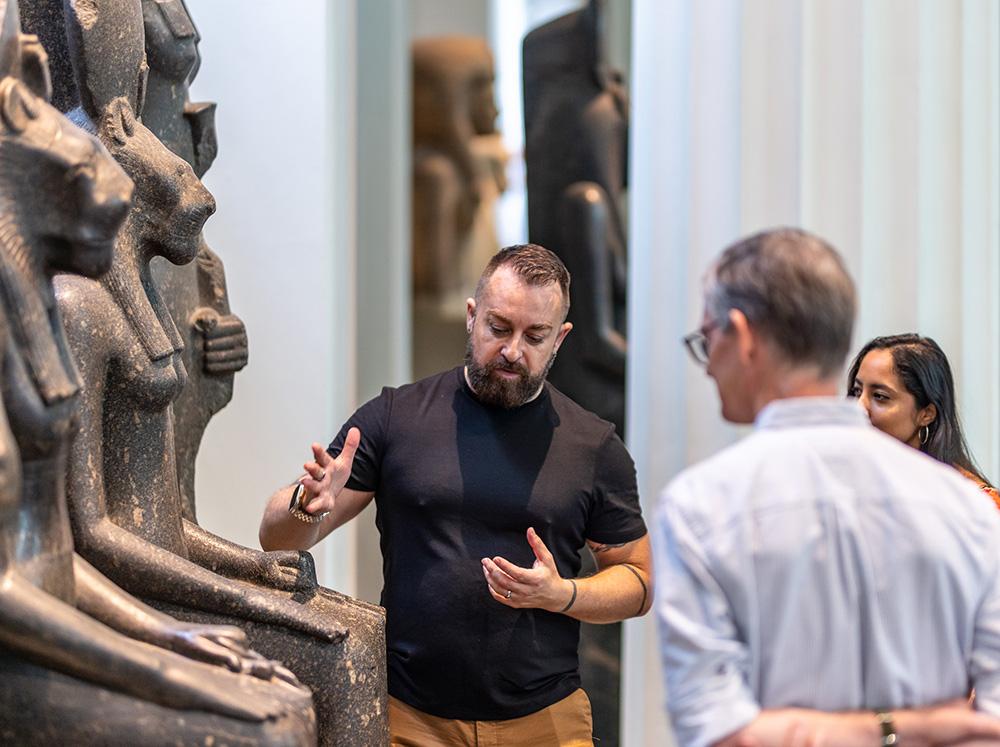 Matthew gestures towards some statues in the Egyptian sculpture gallery, surrounded by visitors