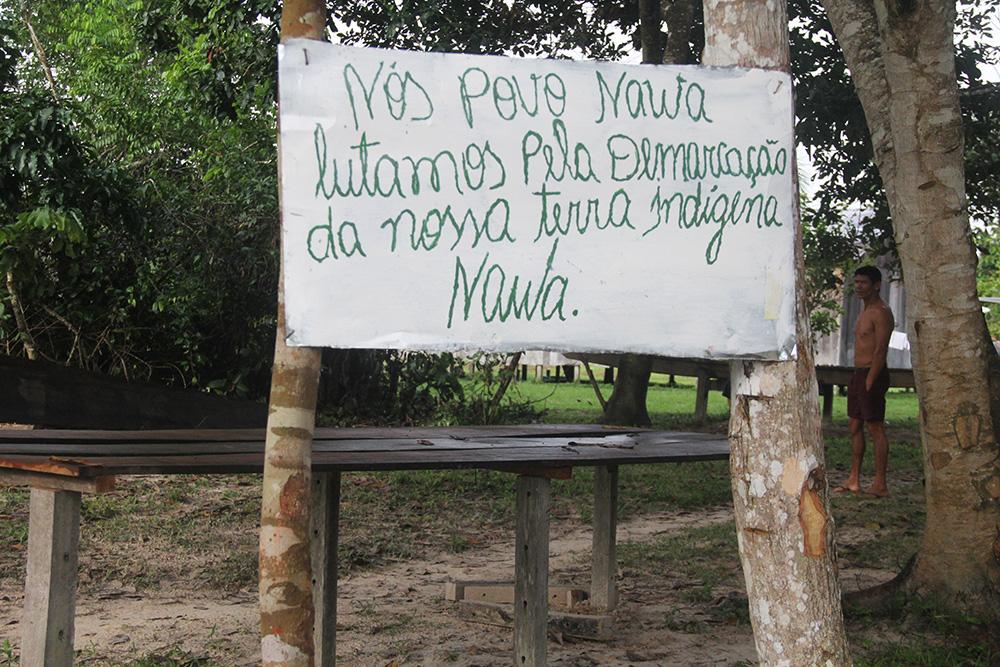 A sign pinned between two trees with writing in Portuguese in cursive script. The words mean: We Nawa people fight for the demarcation of our Nawa Indigenous land
