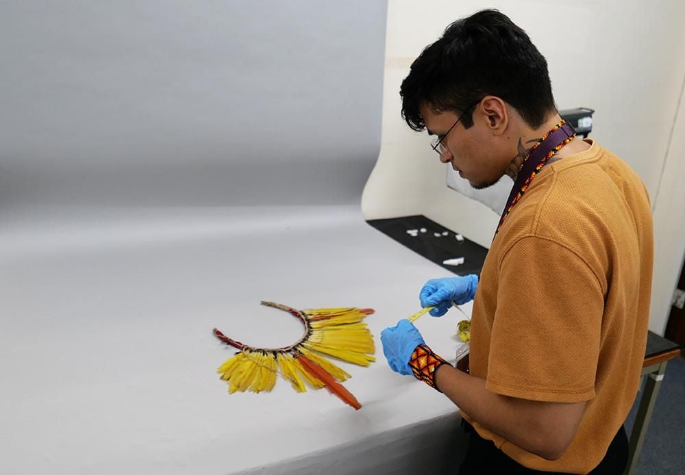 Ykaruni Nawa wears blue protective gloves and holds a tape measure between his hands. In front of him is a headdress made of yellow and red feathers