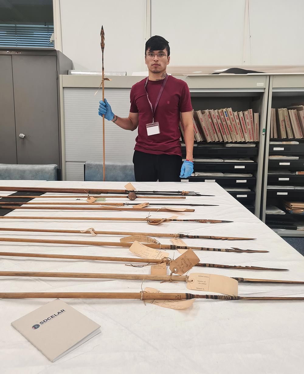 Ykaruni Nawa wears a deep red t-shirt and stands holding a Nawa spear. In front of him is a table of spears with labels tied around them