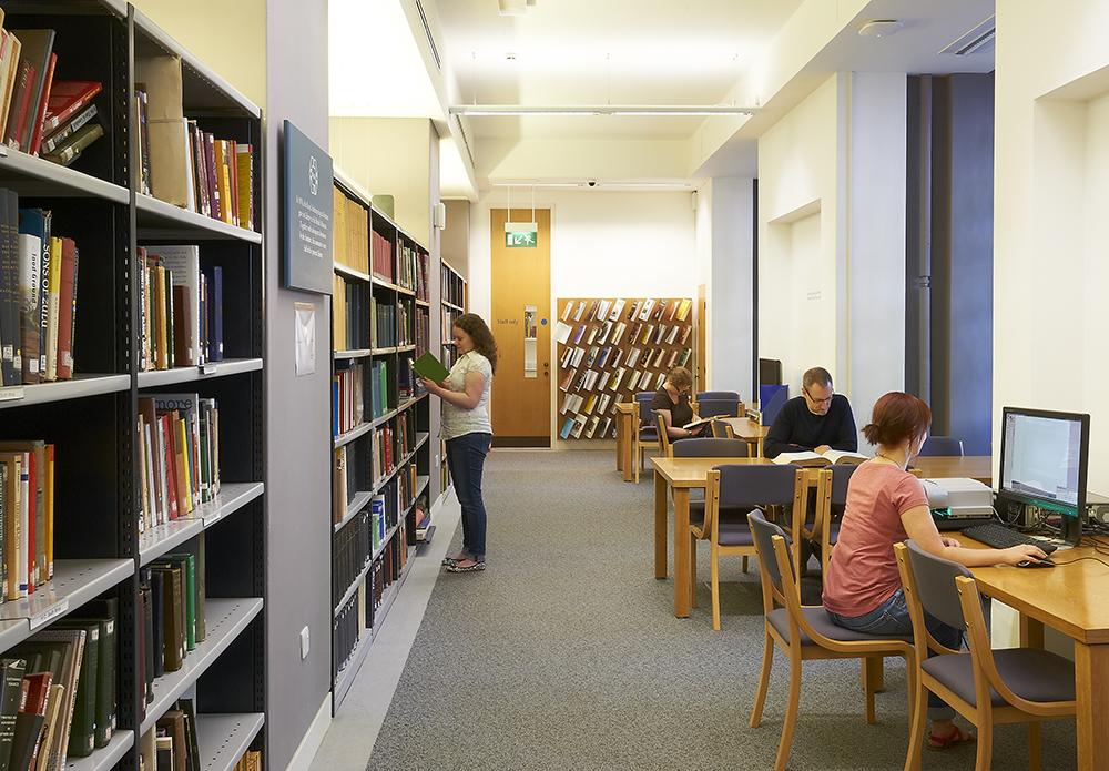 View of a library space with shelves of categorised books lining the left and back walls, with several wooden tables and chairs available on the right.