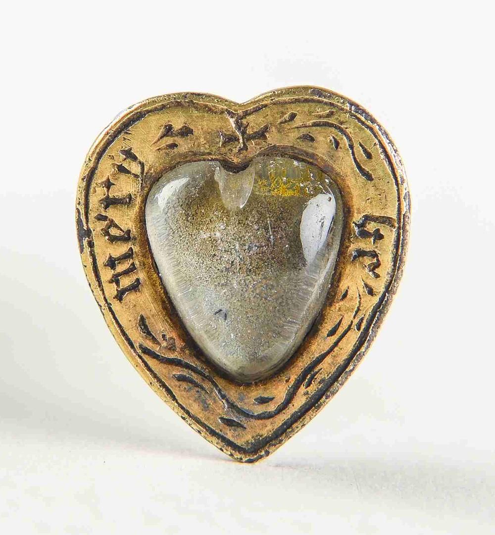 A cap hook in the shape of a heart, with a gold frame and a crystal in the centre. The gold bears the inscription 'be mery'