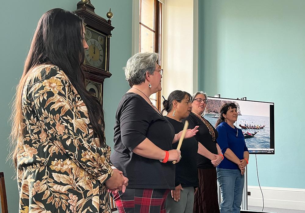Chumash visitors to the British Museum give a presentation on Chumash culture and sing together in a room with blue walls and an old grandfather clock