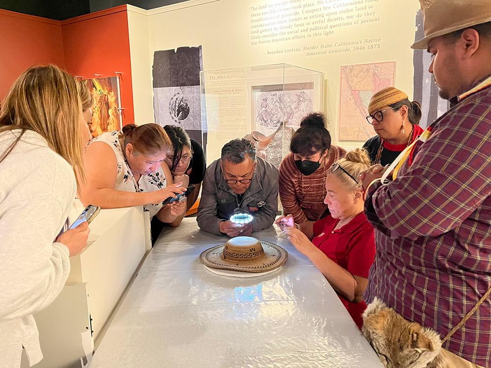 A group of Chumash huddle around the somelelu basketry hat and examine and take photographs of her