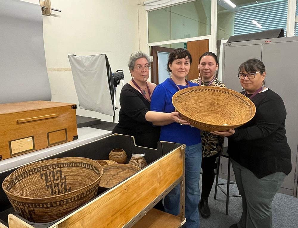 Four Chumash visitors hold a handwoven Chumash basket in their hands and look to camera. They are in a room with other basketry objects and a photographic screen