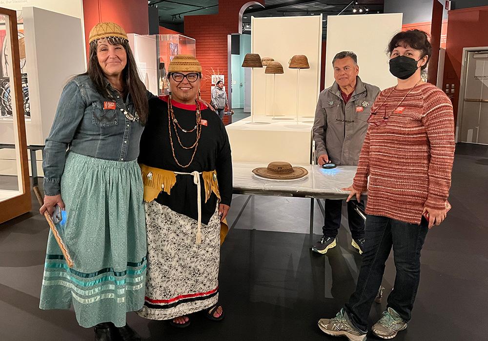 Tima Link stands with other Chumash people in the Autry Museum next to the somelelu which lies on a table. Behind the somelelu are other basketry hats in an open display cabinet