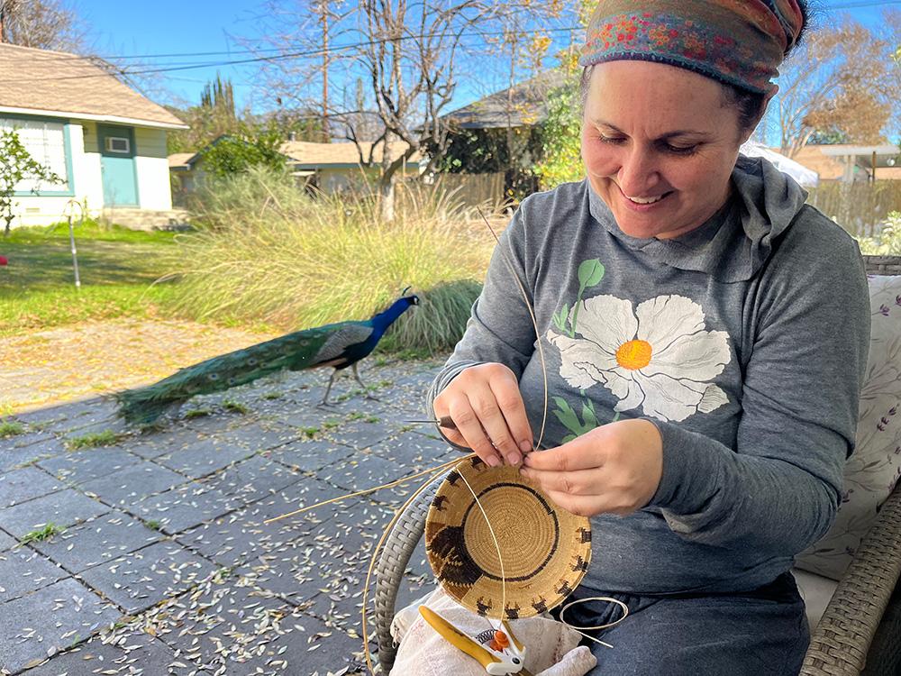 Tima Link wears a grey hoodie with a white flower on it, and weaves a basket in a large garden. Behind her is a peacock, long grass and a white building with a blue door