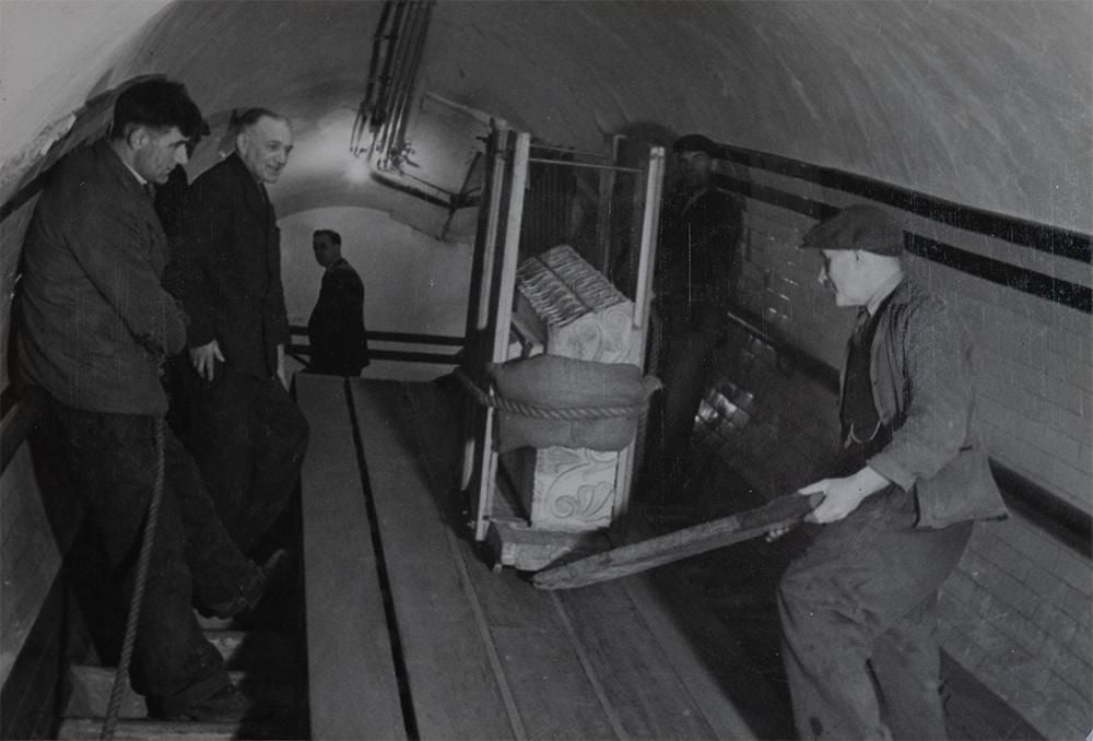Three men lower a stone object down into a tube station during World War II.