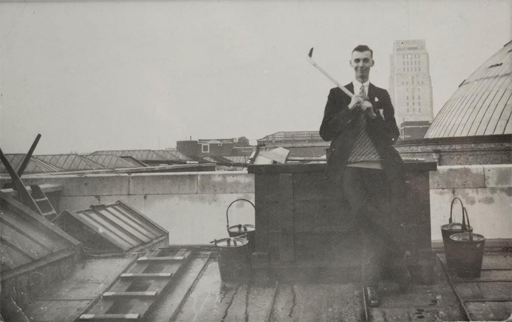 A man in a jacket and tie holds a golf club over his right shoulder and smiles, with his right leg crossed over the left. He stands on the roof of the British Museum, the dome of the Reading Room is visible on the right-hand side and he is surrounded by buckets.