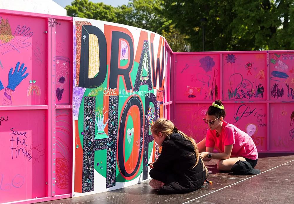 Two young people sat cross-legged outside working on a colourful mural along a pink wall which reads "Draw Hope".