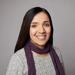 Head and shoulders picture of Rubbena a woman with long, straight dark hair, smiling warmly at the camera. She is wearing a grey knit sweater and a deep purple scarf loosely wrapped around her neck.
