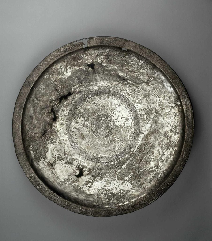 Large shallow silver platter, virtually flat with a flat raised rim and a slight lip, in the centre is a decorated roundel